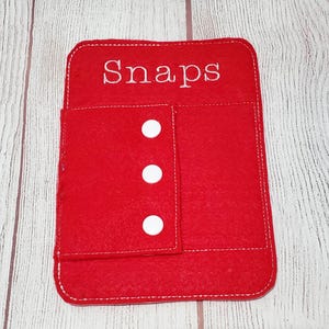 May include: A red felt organiser with white stitching and the word "Snaps" embroidered on the top. It features a flap with three white press studs. The organiser is designed to hold small items.