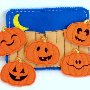 May include: Five felt orange pumpkins with different embroidered facial expressions. The pumpkins are on a blue felt background with a yellow crescent moon.
