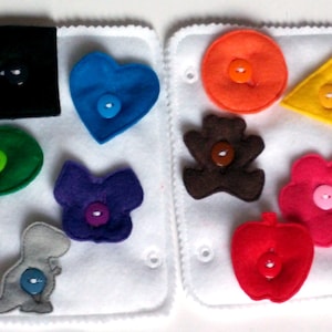 May include: A white felt board with various felt shapes and buttons. The shapes include a heart, a star, a flower, a dinosaur, a butterfly, a circle, a square, and a triangle. Each shape has a button in the center.