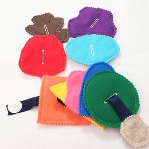 May include: A felt quiet book with various shapes and colors. The shapes include a bear, a cloud, a heart, a circle, and a square. Each shape has a button or a loop for fastening.