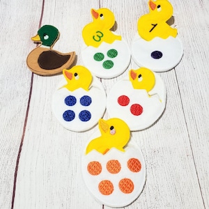 May include: A set of six felt chick and egg counting pieces. Each chick is yellow with a white egg shell. The eggs have different colored dots on them. The numbers 3 and 7 are embroidered on two of the chicks. A brown duck is also included.