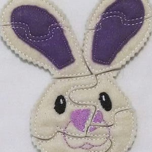 May include: A felt puzzle of a white bunny with purple ears and a pink nose. The puzzle is made up of five pieces.