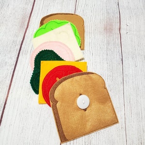 May include: Felt toy sandwich with layers of bread, cheese, tomato, lettuce, and ham. The sandwich is made of felt and has a button in the center.