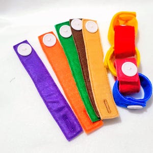 May include: A set of six colorful felt wristbands with white buttons. The wristbands are in the colors purple, orange, green, brown, yellow, and blue.