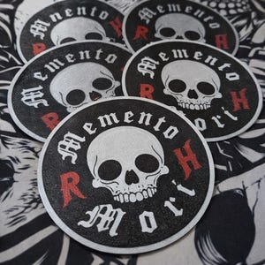 May include: A collection of black and white circular patches with a skull design. Each patch features the words "Memento Mori" in a gothic font, with the letters "R" and "H" in red. The skull is white with black eye sockets and a detailed jawline.