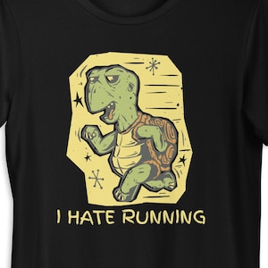 Op de afbeelding: Zwart T-shirt met een cartoon schildpad die rent en er chagrijnig uitziet. De schildpad is groen en bruin, tegen een gele achtergrond met de tekst "I HATE RUNNING". Het T-shirt hangt aan een zwarte hanger.
