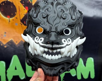 May include: A black and white 3D printed mask with intricate details. The mask features a fierce expression with large teeth and a curled mustache. The eyes are outlined in white and have black pupils. The mask is held in a hand.