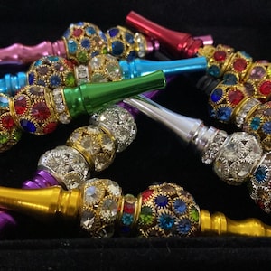 May include: An assortment of colorful hookah mouth tips. The tips are in various colors, including gold, silver, green, blue, and red. Each tip features a decorative, jewel-encrusted ball. The jewels are in a variety of colors, including red, blue, and green.