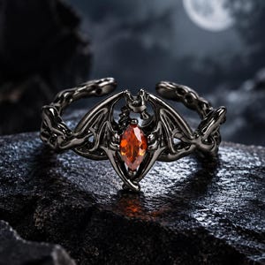 Puede incluir: Un anillo de tono gris oscuro que presenta un diseño de murciélago estilizado con las alas extendidas. El centro del anillo está engastado con una sola piedra preciosa naranja de corte marquesa. La banda tiene un diseño abierto y ajustable con un acabado metálico texturizado.