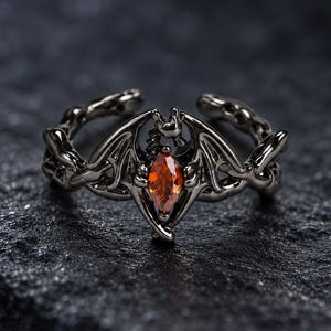 Puede incluir: Un anillo de metal negro con un diseño en forma de murciélago con las alas extendidas. El centro del anillo está engastado con una sola piedra preciosa naranja de talla marquesa. La banda tiene una apariencia texturizada y una abertura trasera.