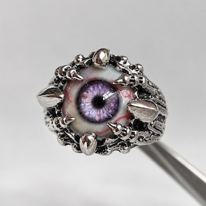 May include: A silver-toned metal ring featuring a central realistic eyeball with a purple iris and red veins. The eye is held in place by a textured, claw-like metal setting that wraps around the circumference of the band.