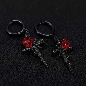 May include: A pair of black hoop earrings with a gothic design. Each earring features a cross pendant with a red rose and a snake detail. The earrings are set against a dark background.
