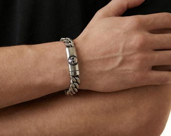 May include: A silver-toned bracelet with a skull accent. The bracelet features a curb chain design and a rectangular clasp with a skull emblem. The bracelet is worn on a person's wrist, with their arms crossed.
