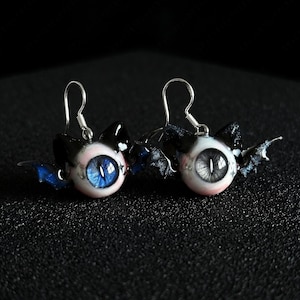 Könnte beinhalten: Ein Paar Ohrhänger mit fledermausförmigen Anhängern mit glänzend schwarzen Ohren und Flügeln. Jeder Ohrring zeigt ein kugelförmiges Auge mit vertikaler Pupille, eines mit leuchtend blauer und das andere mit grauer Iris, an silbernen Haken.