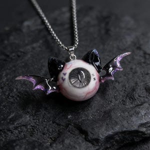 May include: A striking pendant necklace with an eyeball design, complete with black bat wings and purple glitter detailing. The eyeball features a detailed iris and is suspended from a silver chain. The background is dark and textured.