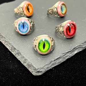 May include: Five silver rings with different colored eye designs. The rings have a detailed, ornate design. The eyes are green, blue, red, orange, and pink.