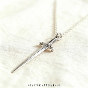 May include: A silver-colored pendant necklace featuring a miniature sword design. The sword has a detailed hilt and a pointed blade, suspended from a delicate chain. The necklace is displayed on a white, textured surface.