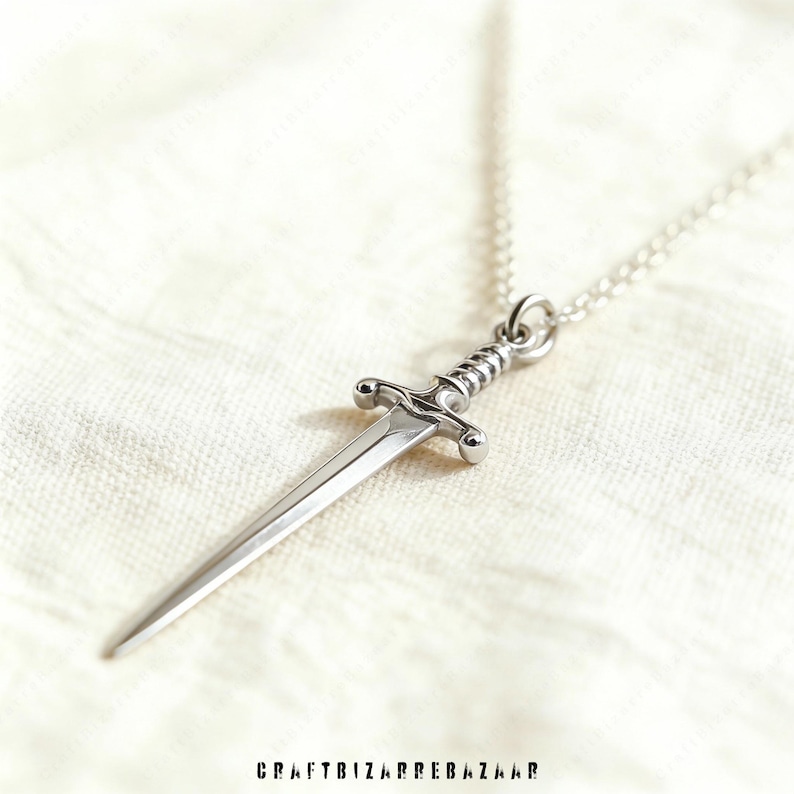 May include: A silver-colored pendant necklace featuring a miniature sword design. The sword has a detailed hilt and a pointed blade, suspended from a delicate chain. The necklace is displayed on a white, textured surface.