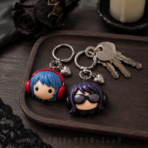 May include: Two character keychains made of clay. One features a figure with blue hair and red headphones. The other features a figure with purple hair and black sunglasses. Both keychains include a silver metal ring and a small heart charm.