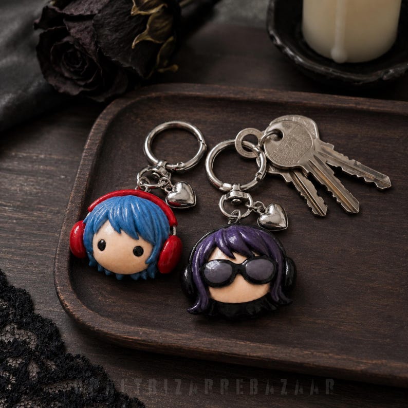 May include: Two character keychains made of clay. One features a figure with blue hair and red headphones. The other features a figure with purple hair and black sunglasses. Both keychains include a silver metal ring and a small heart charm.