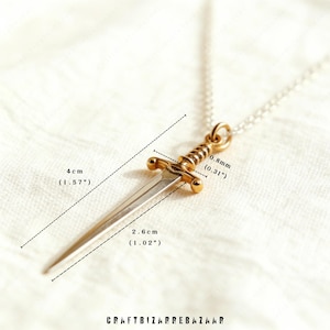 May include: A gold-colored dagger pendant on a silver chain. The dagger measures 1.57 inches long and 1.02 inches wide. The pendant has a detailed hilt and a sharp blade. The necklace is displayed on a white surface. The text "CRAFTBIZARREBAZAAR" is at the bottom.