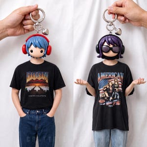 May include: Two keychains featuring stylized figures with large heads, one with blue hair and red headphones, the other with purple hair and black sunglasses. Both figures wear black t-shirts with graphic prints, one reading DOOM MASTER POUPPETS.