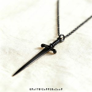 May include: A black dagger pendant necklace. The pendant is a miniature sword with a detailed hilt and a sharp blade. The necklace chain is also black. The background is a textured, off-white fabric. The text "CRAFTBIZARREBAZAAR" is at the bottom.