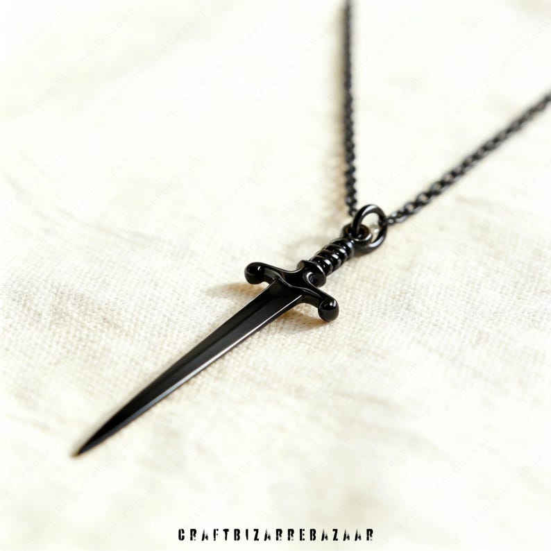 May include: A black dagger pendant necklace. The pendant is a miniature sword with a detailed hilt and a sharp blade. The necklace chain is also black. The background is a textured, off-white fabric. The text "CRAFTBIZARREBAZAAR" is at the bottom.