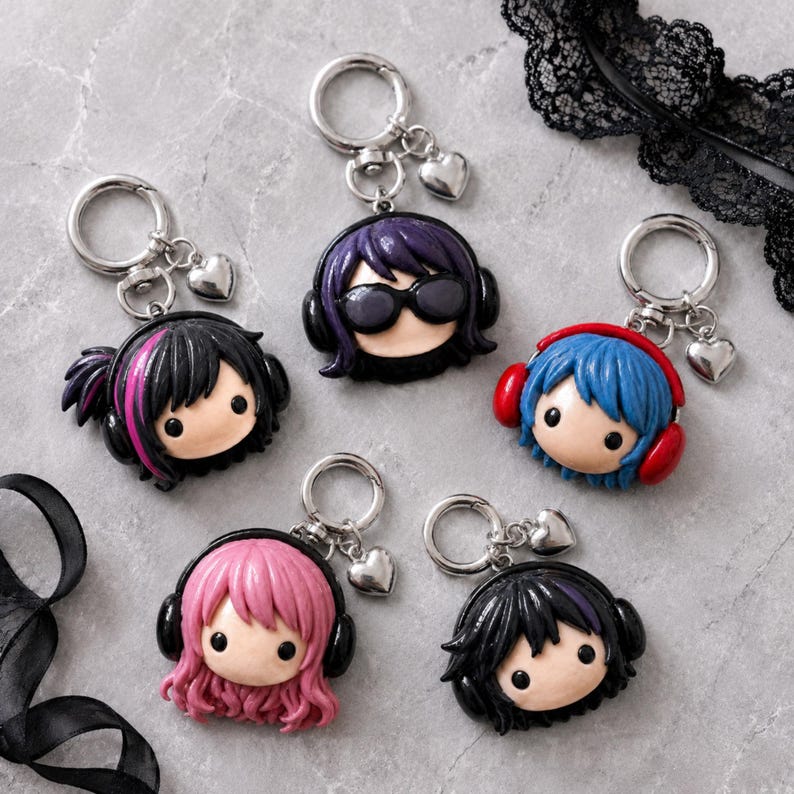 May include: Five keychains featuring chibi-style heads with various hair colors including black with pink streaks, purple, blue, pink, and black. Each head wears headphones and is attached to a silver-toned metal ring with a small heart charm.