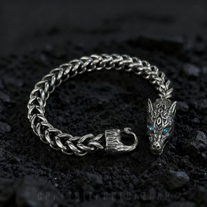 Puede incluir: Una pulsera plateada con un colgante de cabeza de lobo y ojos azules. La pulsera presenta un diseño de cadena tejida y un cierre de gancho. La cabeza de lobo está intrincadamente detallada y la pulsera se muestra sobre una superficie oscura y texturizada.