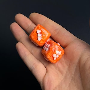 Könnte beinhalten: Zwei orangefarbene Würfel mit weißen Punkten. Die Würfel sind aus einem strukturierten Material gefertigt und haben eine leicht unregelmäßige Form.