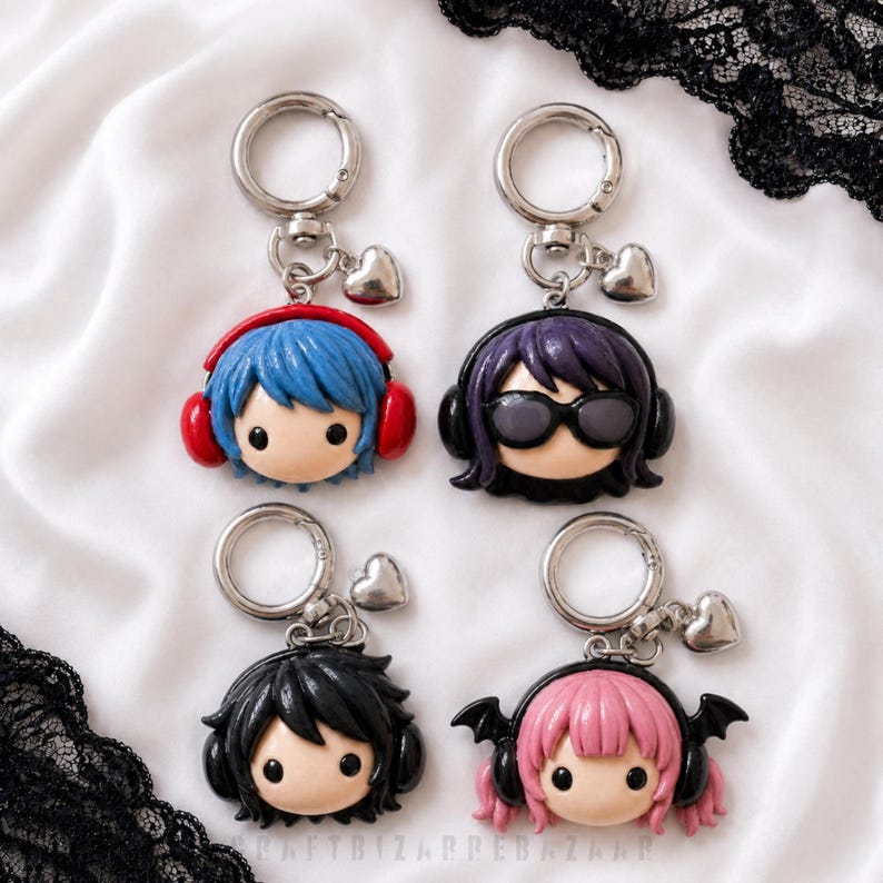 May include: Four keychains featuring chibi-style heads with headphones. One has blue hair and red headphones, one has purple hair and sunglasses, one has black hair and black headphones, and one has pink hair with black bat wings and headphones.