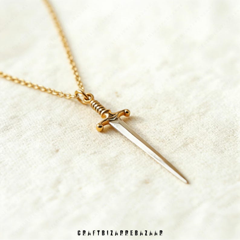 May include: A gold-colored pendant necklace featuring a miniature sword. The sword has a silver-colored blade and a detailed gold hilt. The necklace chain is also gold. The image is set against a white, textured background. The text "CRAFTBIZARREBAZAAR" is at the bottom.