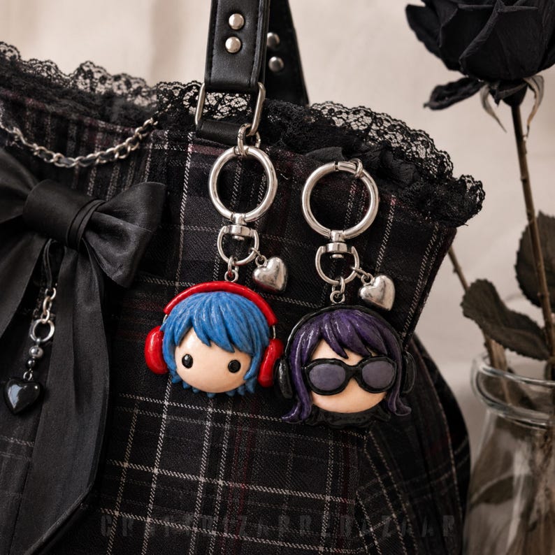 May include: Two character keychains attached to a black plaid bag with a black bow. One keychain features a figure with blue hair and red headphones, the other a figure with purple hair and black sunglasses. Both have silver heart charms.