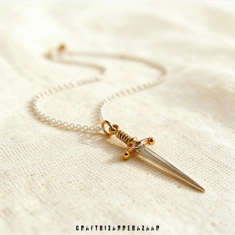 May include: A silver chain necklace with a gold-colored dagger pendant. The dagger has a silver blade and a gold-colored hilt with intricate detailing. The necklace is displayed on a cream-colored fabric. The text "CRAFTBIZARREBAZAAR" is at the bottom.