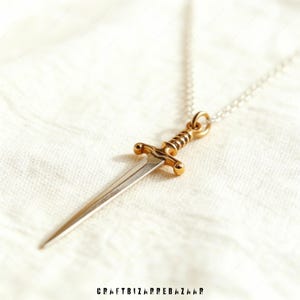 May include: A gold and silver dagger pendant necklace. The dagger has a gold hilt and a silver blade. The necklace is on a silver chain. The pendant is displayed on a white, textured surface. The text "CRAFTBIZARREBAZAAR" is at the bottom.