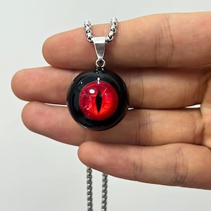 May include: A silver chain necklace with a black and red eye pendant. The eye is red with a black pupil and a black outline.