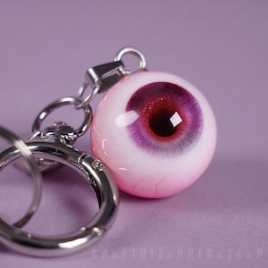 May include: A novelty keychain featuring a realistic eyeball pendant. The pendant has a pink sclera, a deep red iris, and a black pupil. The keychain includes a silver-colored clasp and ring.