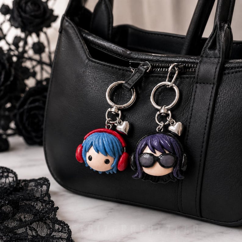 May include: Two character keychains attached to a black handbag. One features a figure with blue hair and red headphones, the other a figure with purple hair and black sunglasses. Both charms include a silver ring and a small heart-shaped charm.