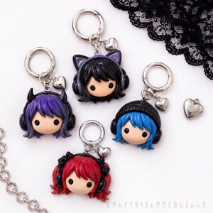 May include: Four keychains featuring chibi-style heads with colorful hair, black headphones, and small silver heart charms. The characters have purple, black, blue, and red hair, with one sporting small horns and another wearing a black beanie.