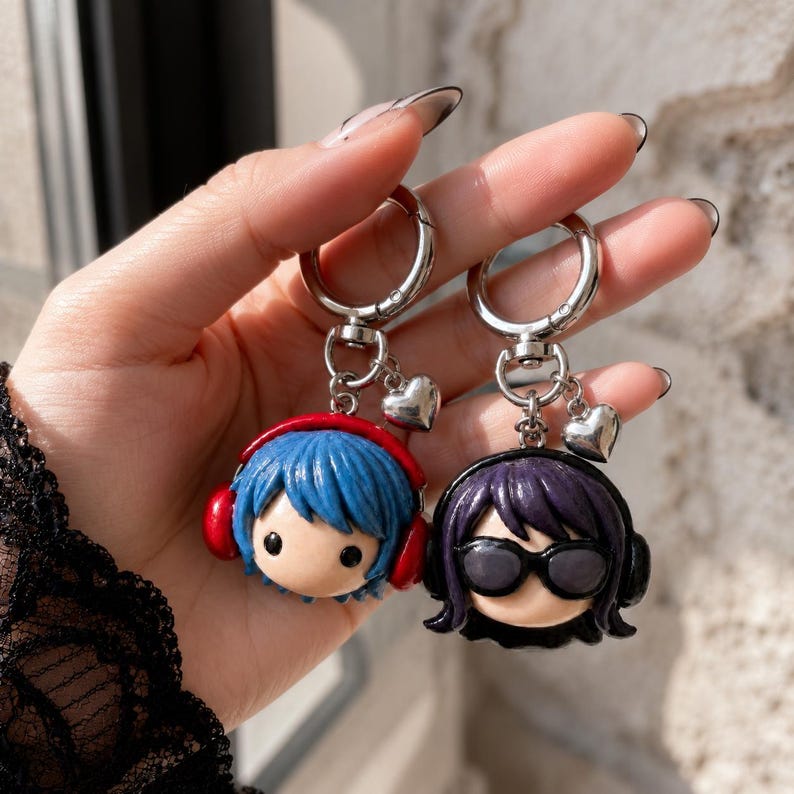 May include: Two character keychains featuring sculpted heads with silver-toned metal rings and small heart charms. One figure has blue hair and red headphones, while the other has dark purple hair and black sunglasses with matching black headphones.