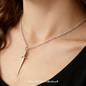 May include: A silver chain necklace with a small dagger pendant. The dagger has a gold-colored hilt and a silver blade. The necklace is displayed on a person wearing a black top. The text "CRAFTBIZBAZAAR" is visible at the bottom.
