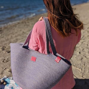May include: A large, light purple and white woven tote bag with red leather accents. The bag has two long straps and is made of a thick, textured material.