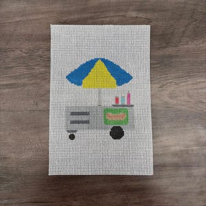 May include: A needlepoint canvas featuring a colorful illustration of a food cart. The cart has a blue and yellow umbrella, a green body, and black wheels. The canvas is white with a grid pattern, and the cart is filled with colorful items.