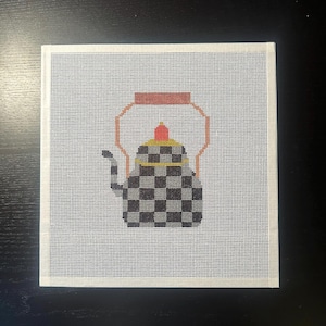 May include: Needlepoint canvas featuring a checkered teapot design. The teapot has a brown handle, a red and yellow top, and a black and gray checkered body. The canvas is square and has a white border.