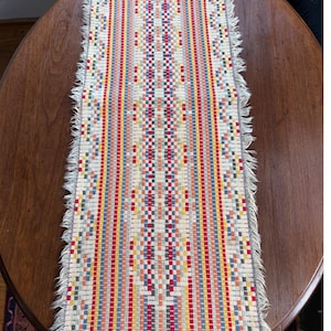 May include: A rectangular woven table runner with a colorful striped pattern. The design incorporates red, blue, yellow, and white vertical stripes with a central diamond pattern. The edges are finished with a fringe.