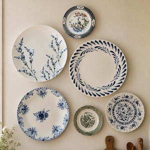 May include: A collection of six decorative ceramic plates mounted on a wall, featuring various blue and white floral, botanical, and landscape patterns. Below, a wooden bowl holds garlic bulbs next to a ceramic vase and kitchen utensils.