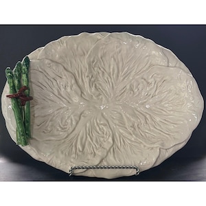 May include: An oval ceramic serving platter shaped like a large cabbage leaf in an off-white color. On the left side, three green ceramic asparagus stalks are bundled together with a brown ceramic tie, creating a raised, textured detail.