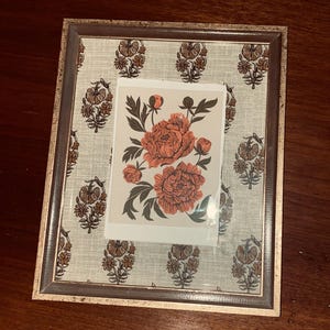 May include: A rectangular picture frame with a floral print background and a floral print art piece. The frame has a gold and brown finish. The art piece features coral-colored peonies with green leaves.