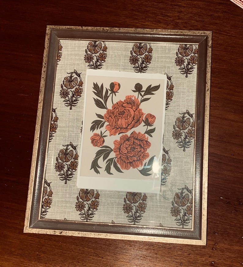 May include: A rectangular picture frame with a floral print background and a floral print art piece. The frame has a gold and brown finish. The art piece features coral-colored peonies with green leaves.